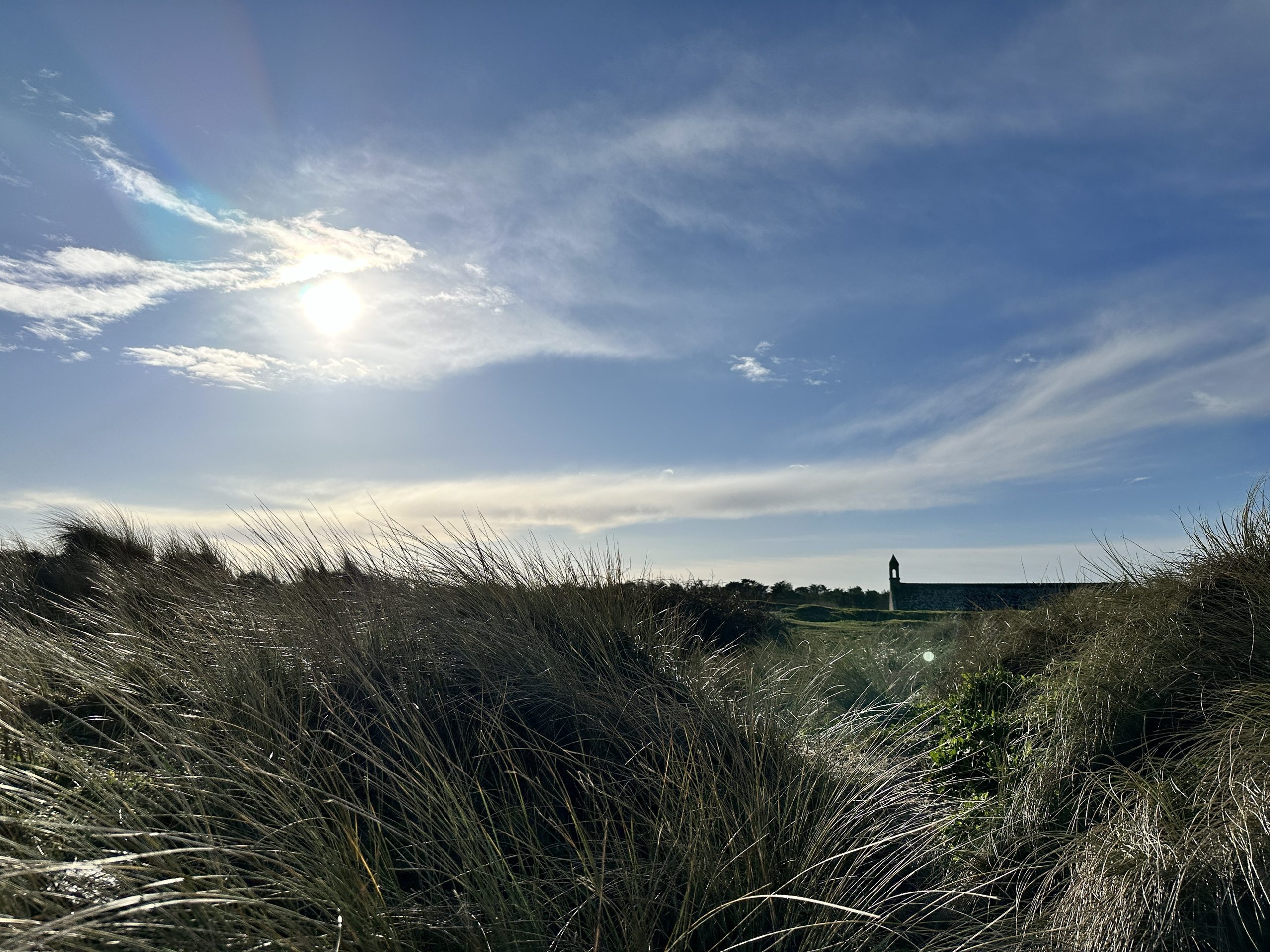 Marram grass on coastal dunes under a blue sky, a stone chapel with a small bell tower on the horizon